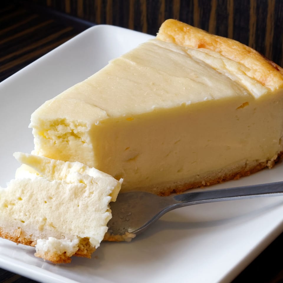 Slice of creamy cheesecake with a golden crust on a white plate, accompanied by a fork and separate bite-sized piece.