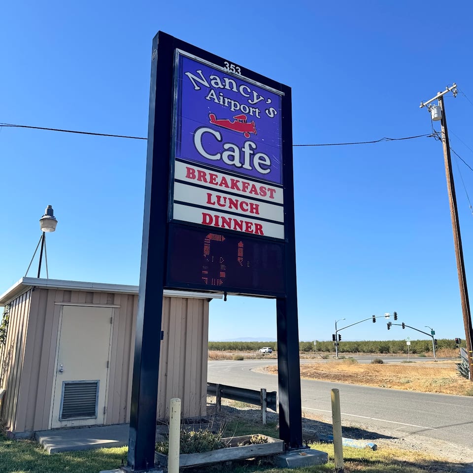 Roadside sign for Nancy Airport's Cafe advertising breakfast, lunch, and dinner beside a rural intersection.