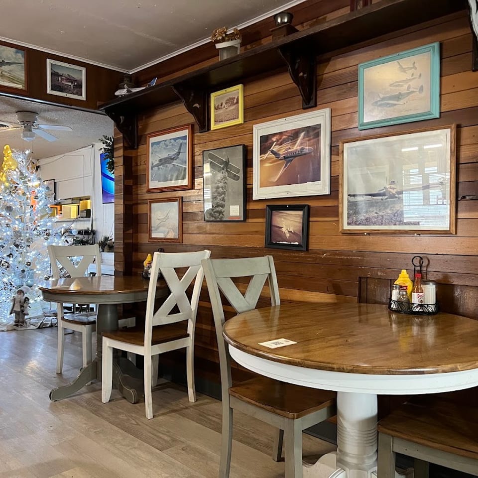 Cozy restaurant dining area with wooden walls, aviation artwork, round tables, white chairs, and a decorated Christmas tree.