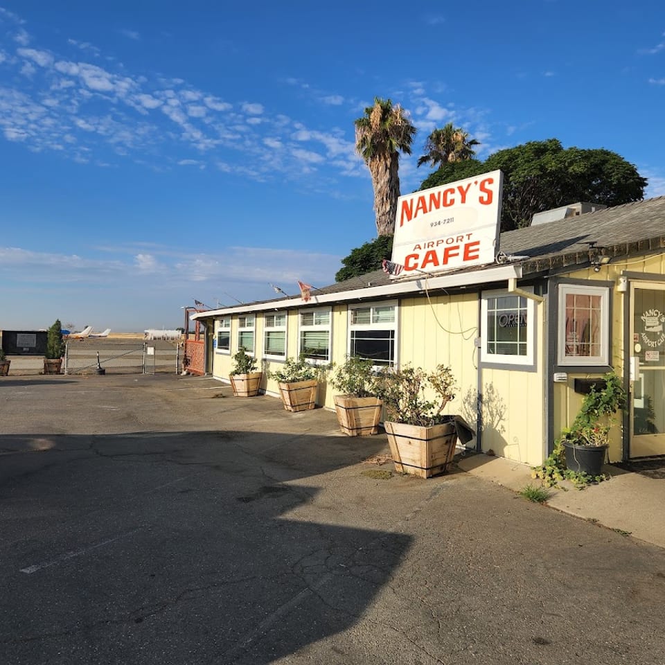 Nancy’s Airport Cafe exterior with yellow siding, rooftop sign, palm trees, potted plants, and airplanes visible beyond the parking lot.