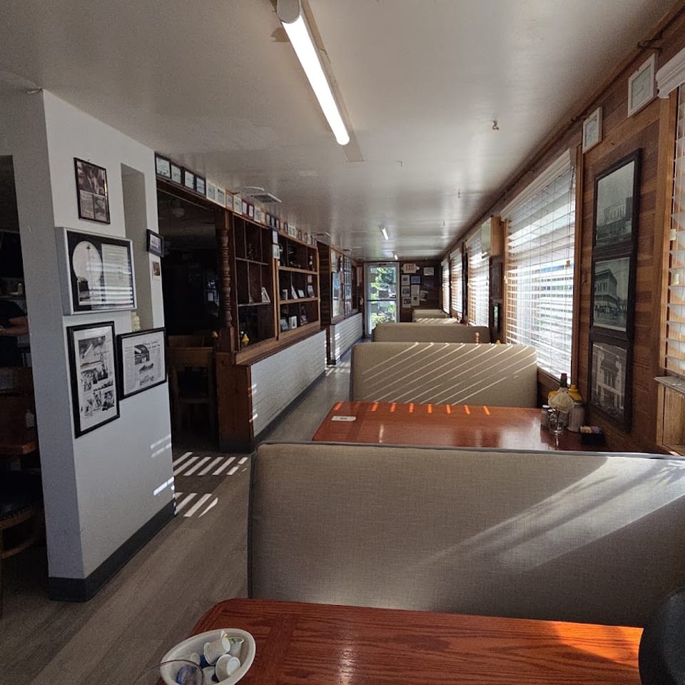 Sunlit restaurant dining area with wood-paneled walls, booth seating, framed pictures, and large windows with blinds.