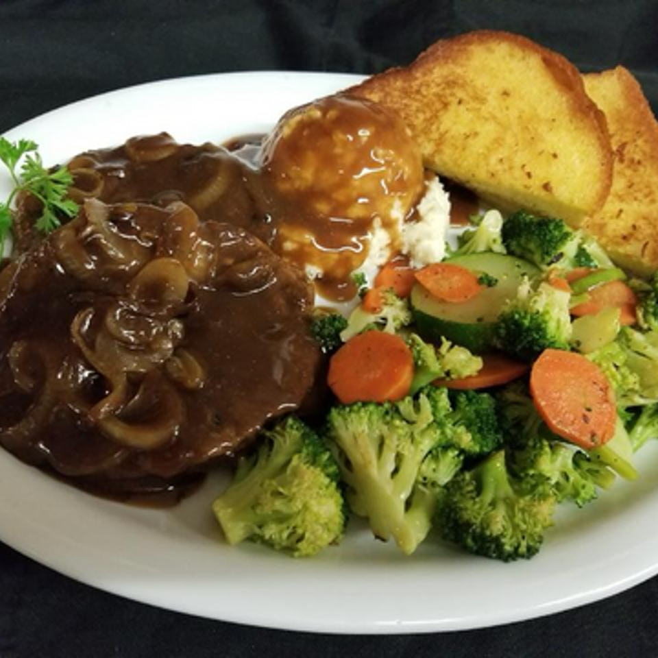 Salisbury steak topped with mushroom gravy, mashed potatoes, toasted bread, and steamed broccoli, carrots, and zucchini on a white plate.