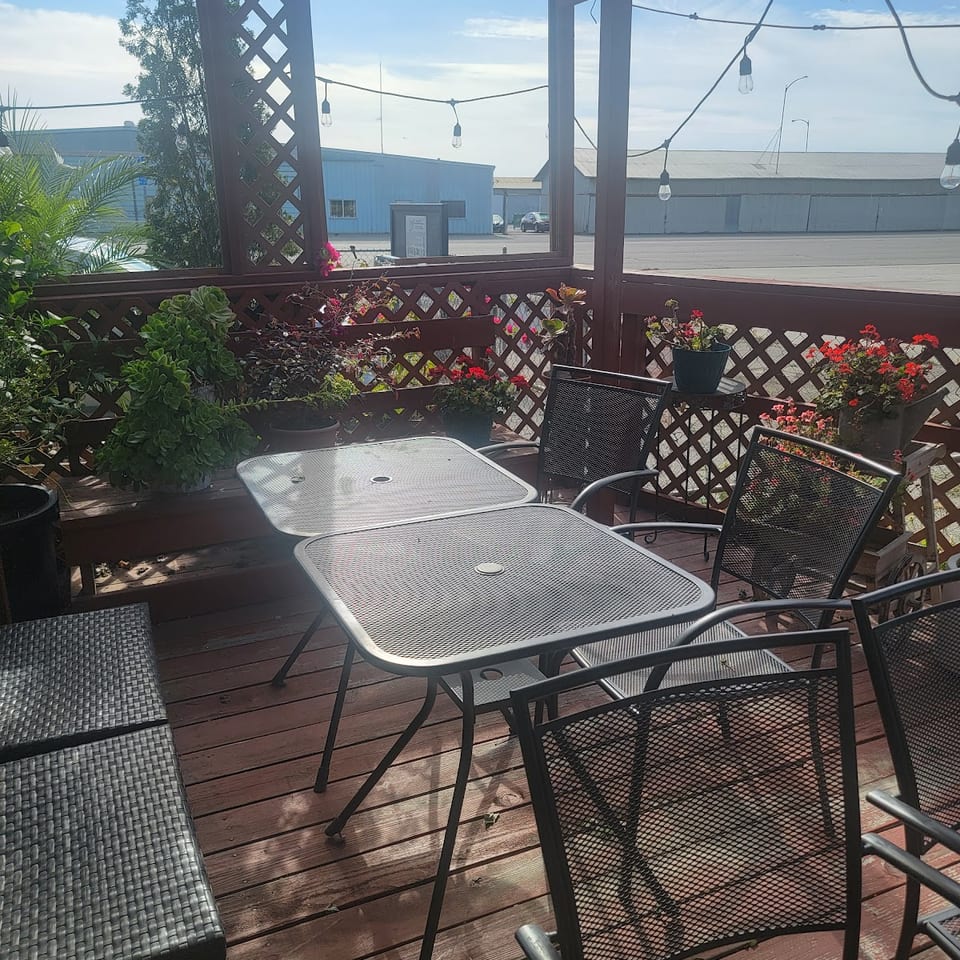 Outdoor restaurant patio with metal tables, chairs, potted flowers, lattice railings, and hanging string lights overlooking industrial buildings.