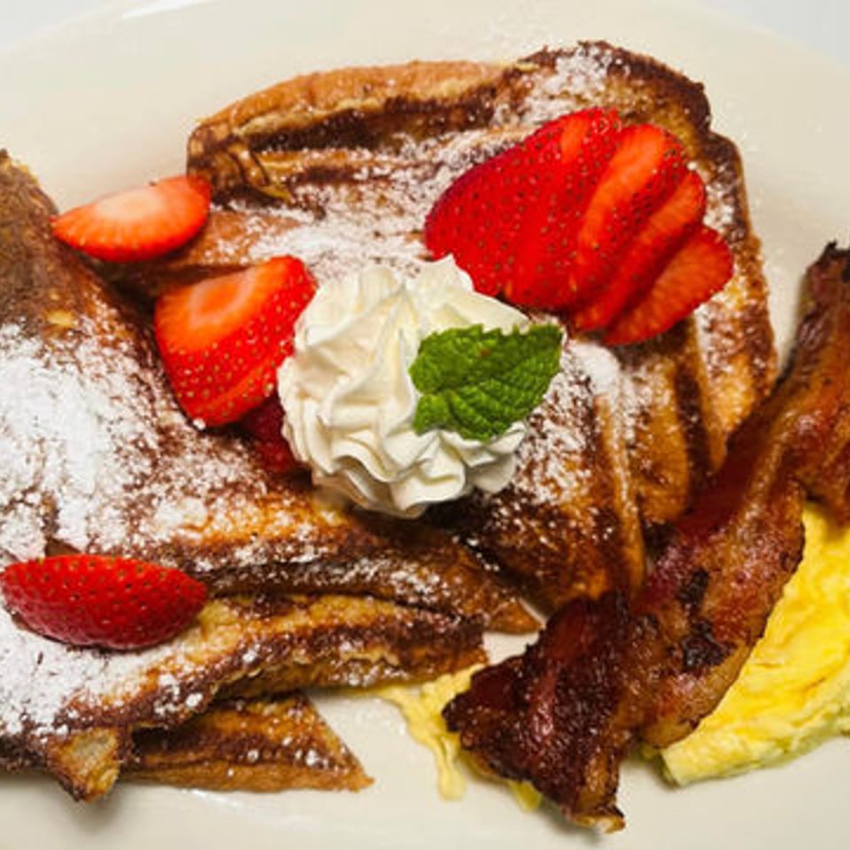 French toast topped with powdered sugar, strawberries, whipped cream, and mint, served with bacon and scrambled eggs.